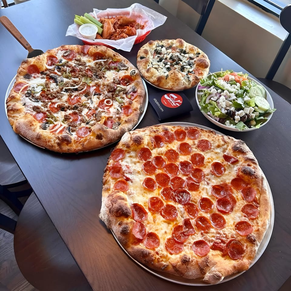 Two large pizzas, a smaller pizza, chicken wings, and a salad on a dark wooden table.