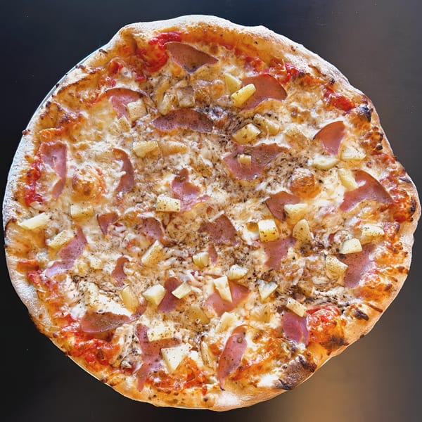 A whole Sid's Pizza with ham, pineapple, and cheese on a dark surface.