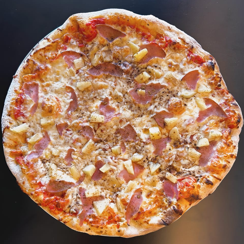 A whole Sid's Pizza with ham, pineapple, and cheese on a dark surface.