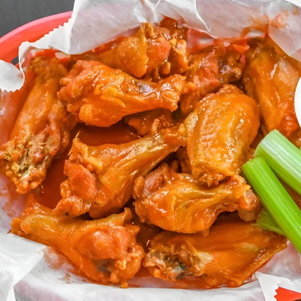 A basket of Sid's Pizza's buffalo wings, coated in orange sauce, with celery sticks.