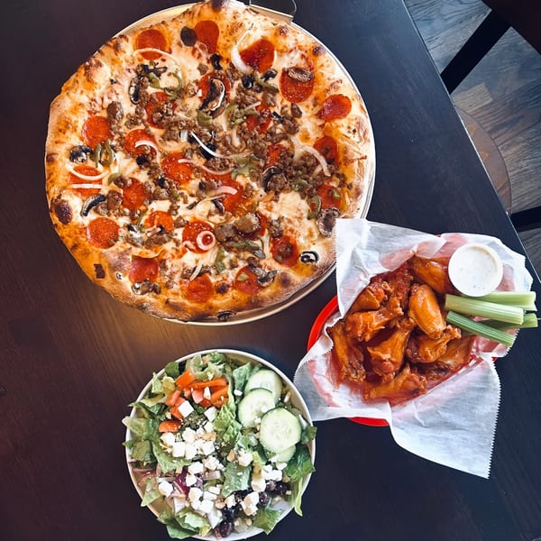 Sid's Pizza Alpharetta's pepperoni, sausage, and veggie pizza, wings, and salad on a dark table.