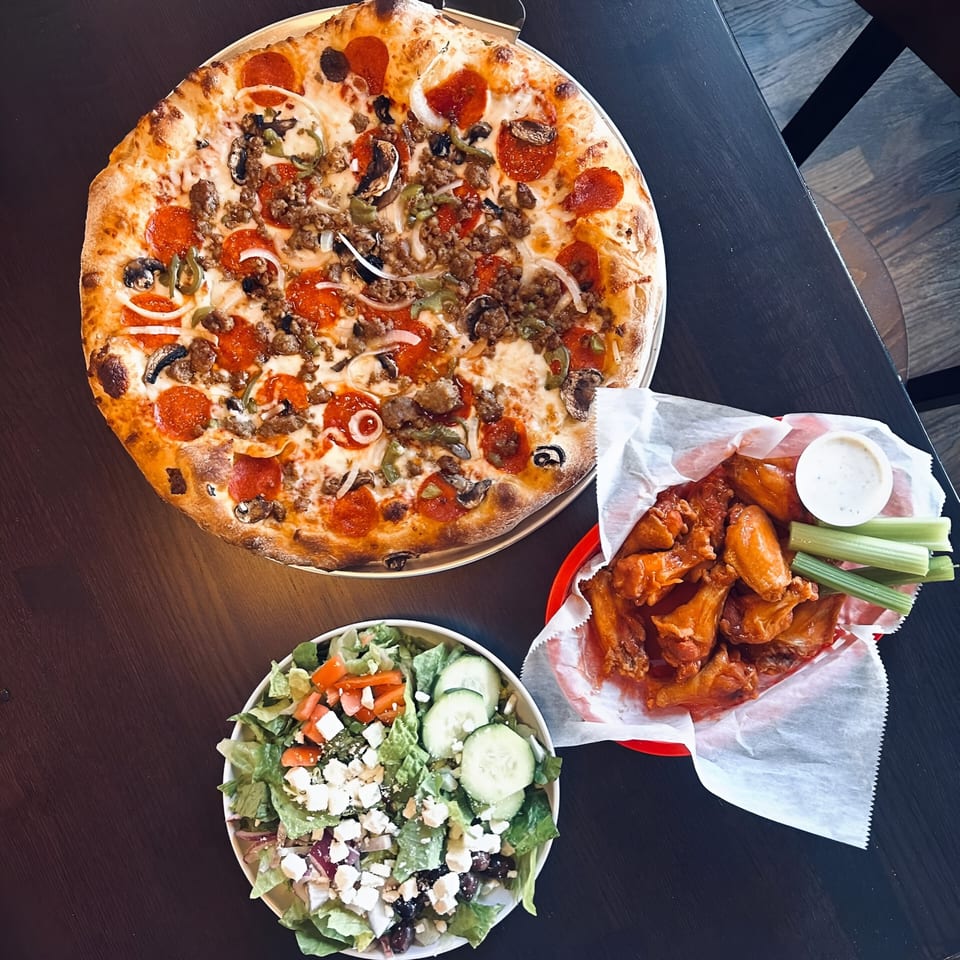 Sid's Pizza Alpharetta's pepperoni, sausage, and veggie pizza, wings, and salad on a dark table.